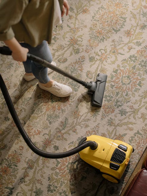 A person holding a yellow portable carpet cleaning machine with a black vent and control panel is performing surface cleaning on an ornate, multicolored area rug with floral patterns. The rug is situa