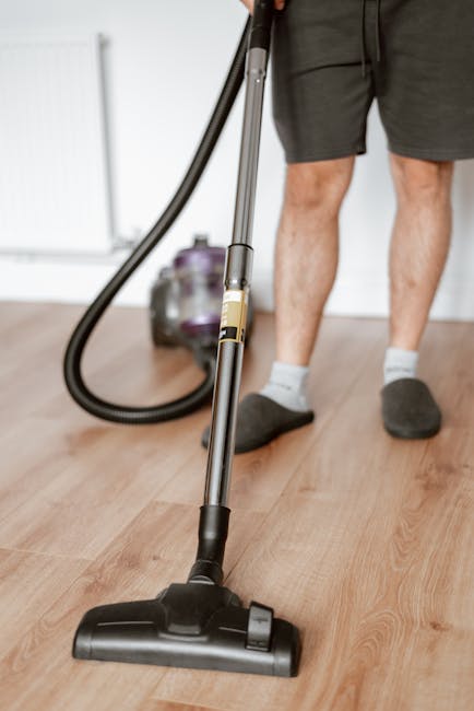 A woman vacuuming a patterned area rug in a living room with wooden flooring visible along the edges. The rug displays a floral design in muted colors. She is using a yellow canister vacuum cleaner wi