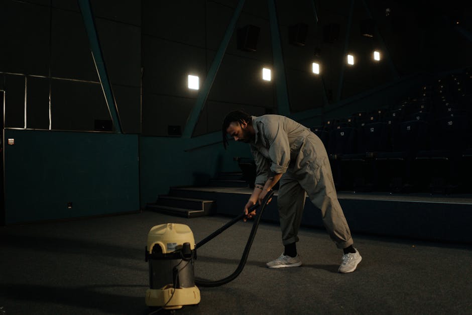 Close-up of a person vacuuming a patterned, beige and green carpet in a room, with the vacuum cleaner's black head and hose visible. The individual, partially visible, is wearing dark pants and white 